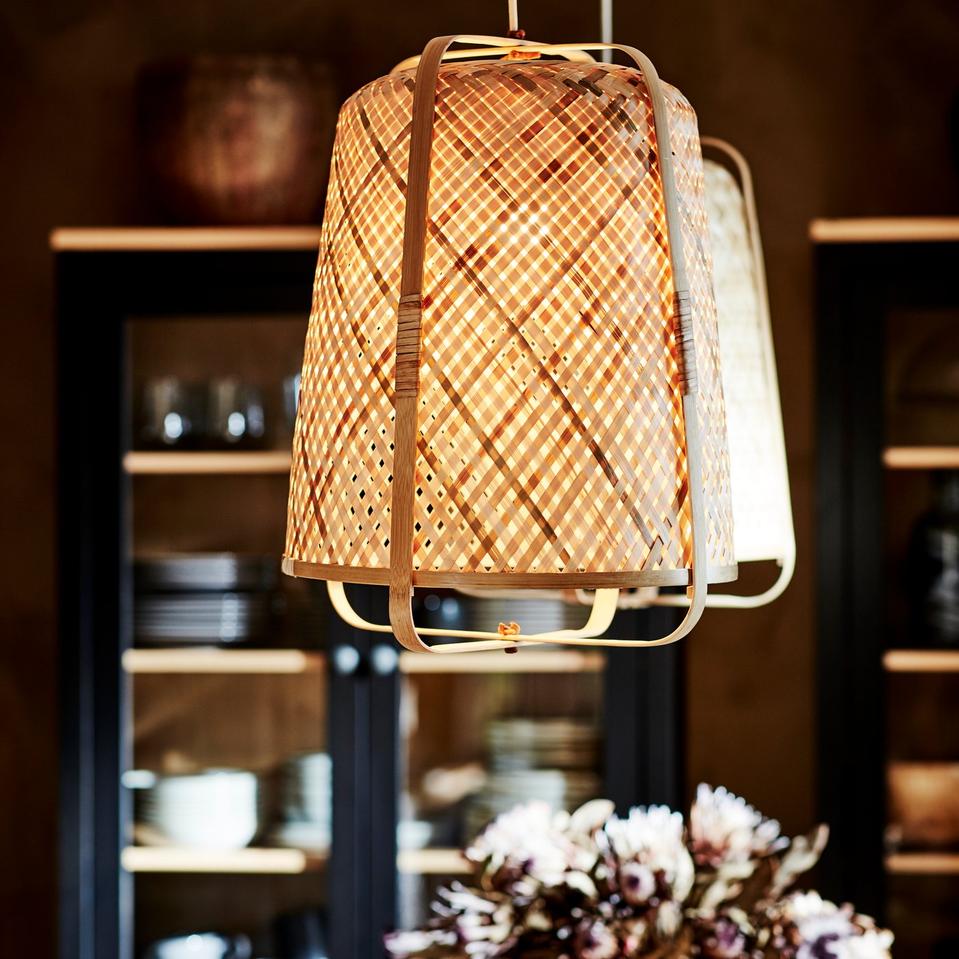 Two KNIXHULT pendant lamps in a dining room. In the background are a bunch of flowers and a shelving unit full of tableware.