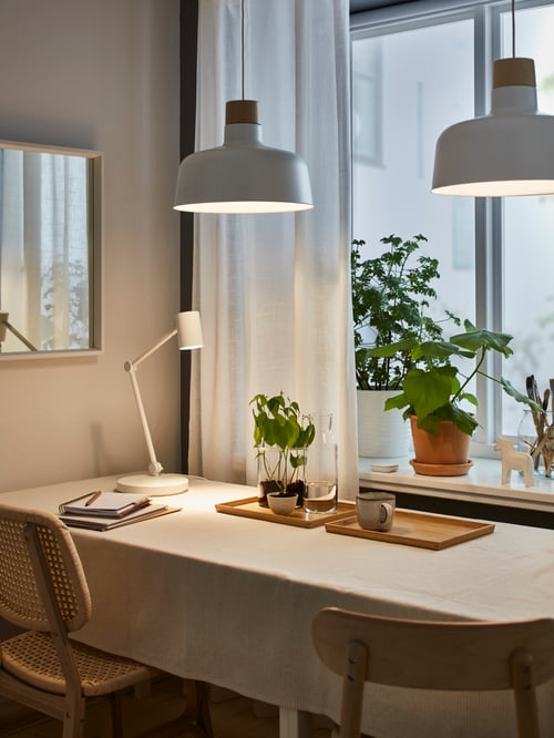 Dining room with 2 white pendant lamps made of metal placed above a rectangular table supporting 2 trays and a mug.