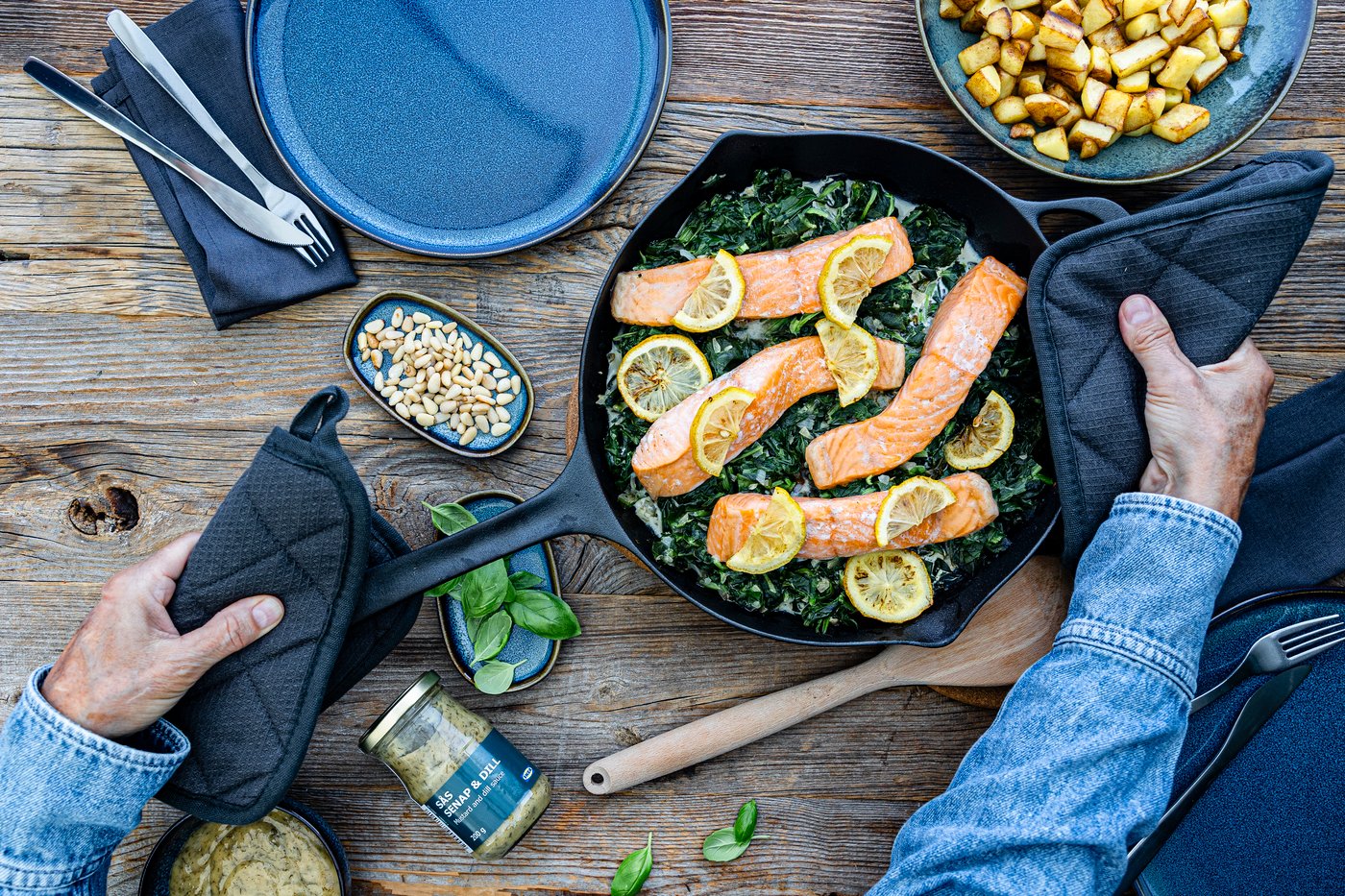 Eine schwarze Pfanne, in der sich Spinat, Lachsfilets und Zitronen befinden. Um die Pfanne stehen Kochutensilien.
