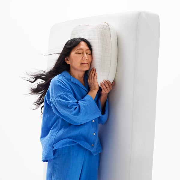 A woman stands up against a bed mattress, leaning her head into a KLUBBSPORRE ergonomic cooling pillow.