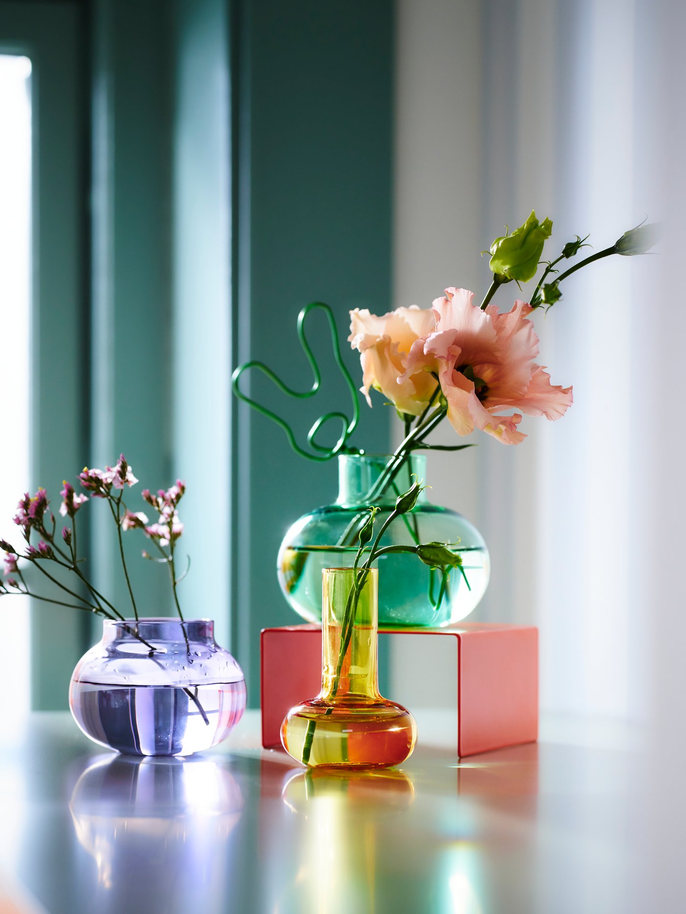 Drei bunte TJÄRLEK Vasen stehen mit einzelnen Blumen dekoriert auf einem Tisch.