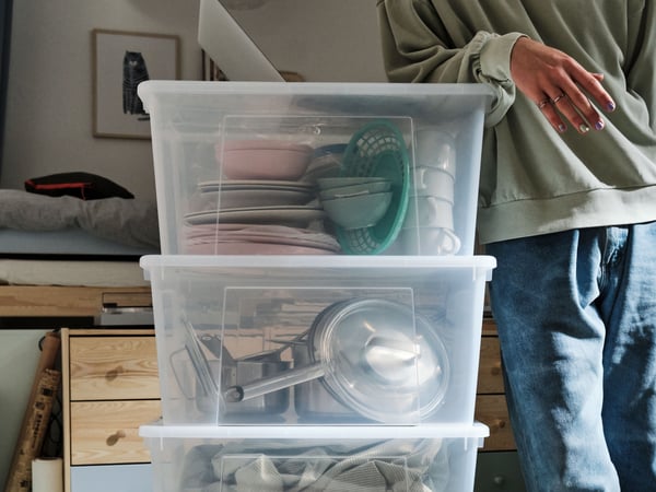 Tre genomskinliga SAMLA förvaringslådor staplade på varandra med köksartiklar, kastruller och textilier. En person står bredvid, och en byrå samt tavlor syns i bakgrunden.
