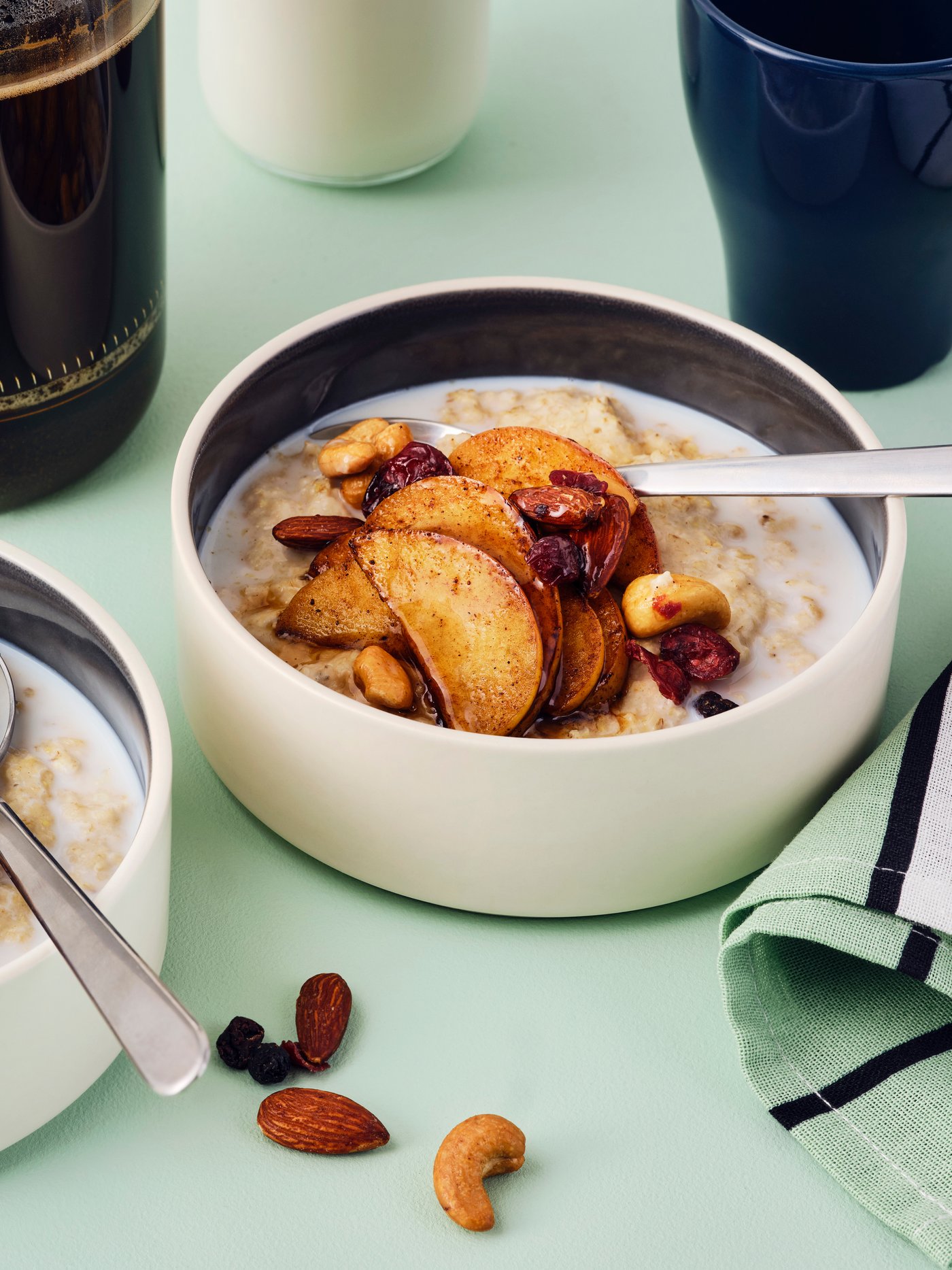 Two bowls of oatmeal with cinnamon-fried apples and MUNSBIT mix roast nuts/berries, and coffee and milk in the background.