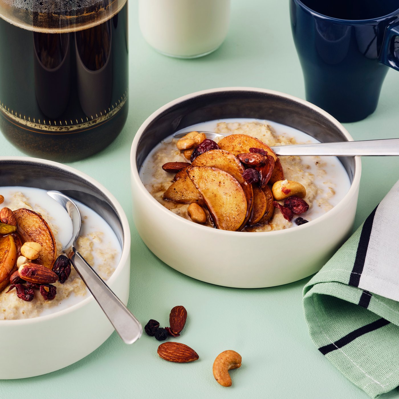 Two bowls of porridge with cinnamon-fried apples and MUNSBIT mix roast nuts/berries, and coffee and milk in the background.