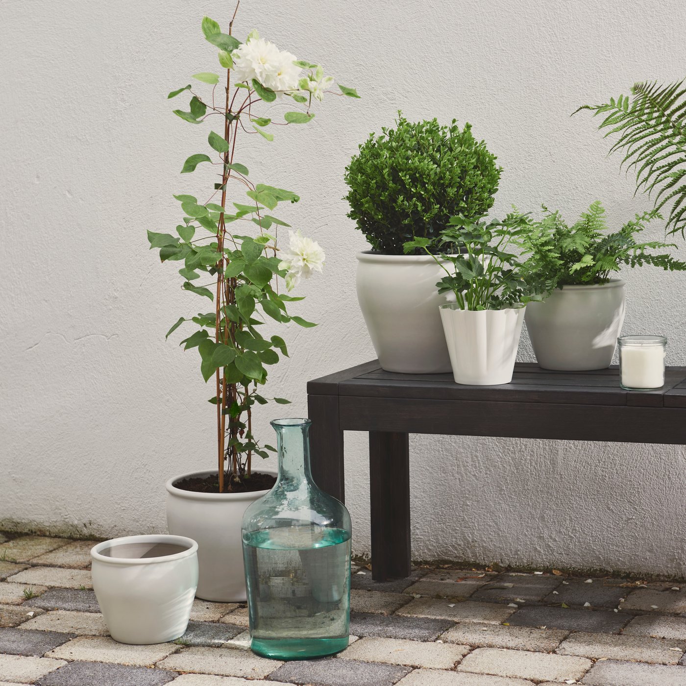 A patio area features a white AMBRARÖNN plant pot, surrounded by lush green plants and outdoor decor.