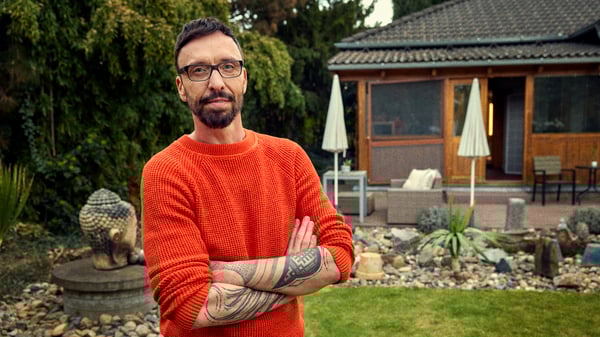 Daniel standing outside in the garden in front of his home, with his arms crossed wearing a red sweater.