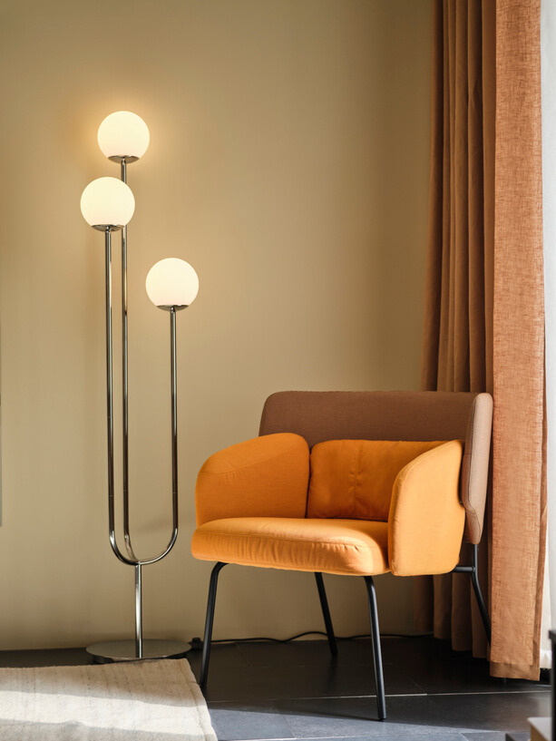 En SIMRISHAMN golvlampa i kromplätering med opalvitt glas bredvid en orange tygfåtölj.
