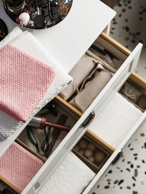 Un moble de lavabo obert amb calaixos TÄNNFORSEN que contenen tovalloles de color blanc i rosa, un assecador i diversos accessoris organitzats.