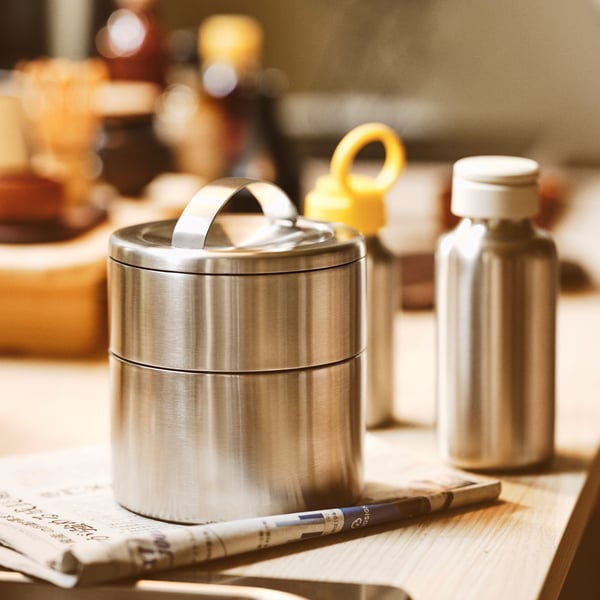 A worktop with an FÖRSKAFFA insulated tiffin box on a newspaper, plus a stainless steel/beige ENKELSPÅRIG water bottle.