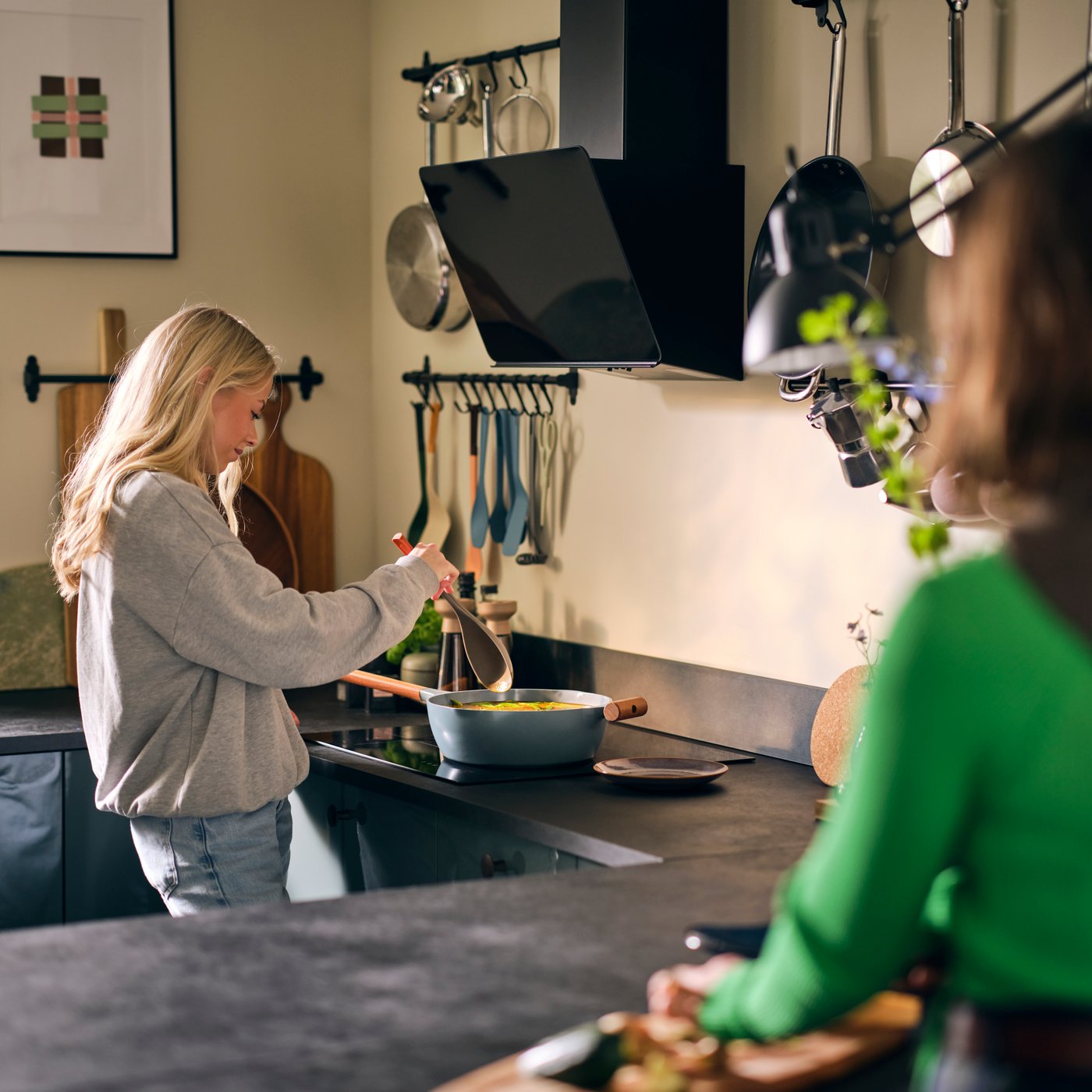 Une cuisine avec un extracteur réglable STACKBO noir, deux femmes, l'une d'entre elles cuisine dans une casserole sur la plaque de cuisson.