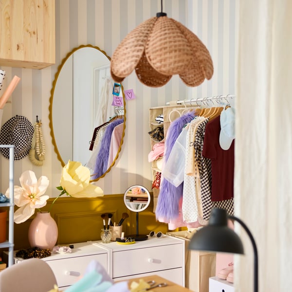 A dressing area with a dark yellow DAGGKAPRIFOL mirror reflecting light, surrounded by clothes on hangers and a flower vase.