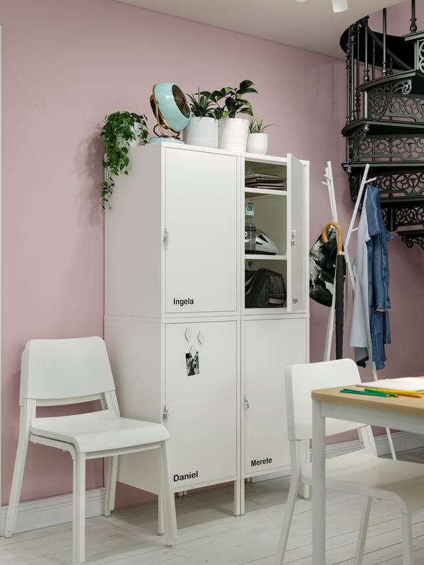White storage combinations marked with names of colleagues, white chairs, a dining table, potted plants.