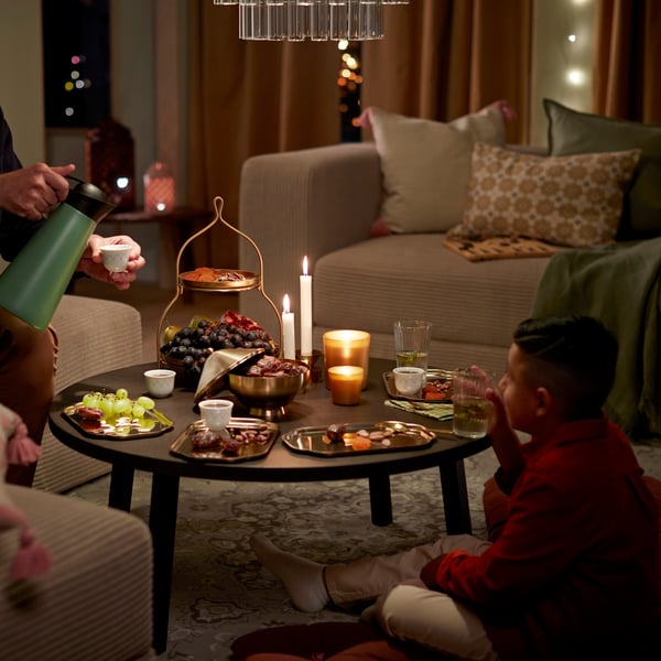 A gold-coloured GOKVÄLLÅ two-tier serving stand displays a variety of snacks on a low table in a cosy living room setting.