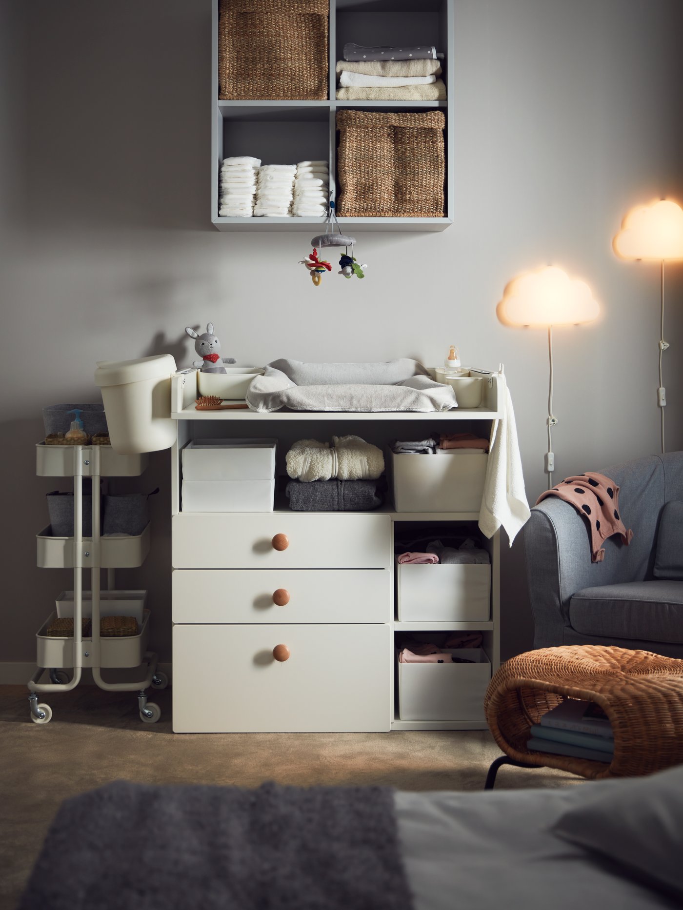 A white SMÅSTAD changing table with 3 drawers, a natural material footstool, a storage trolley and a grey armchair in a cosy room.