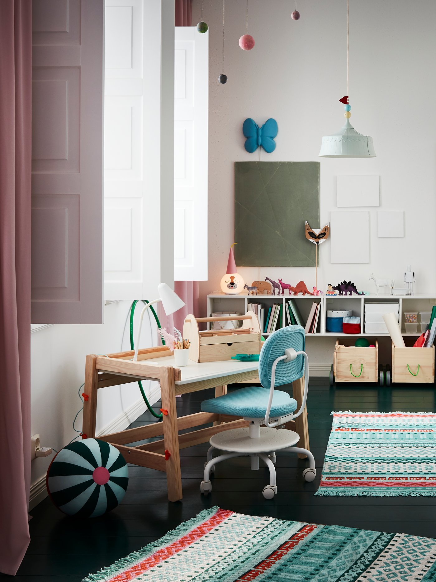 Ein Kinderzimmer im skandinavischen Einrichtungsstil mit kleinem Schreibtisch und Stuhl. An der hinteren Wand befindet sich eine Ablagefläche mit Büchern und Spielzeug. Auf dem Boden liegen Teppiche und Kuscheltiere im Boho Stil.