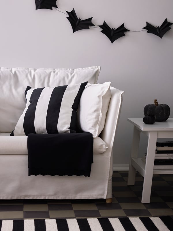 Ein gestreifter weiß/schwarzer HÖSTAGILLE Kissenbezug im Wohnzimmer auf einem weißen Sofa. An der Wand kann man Halloween Deko sehen.