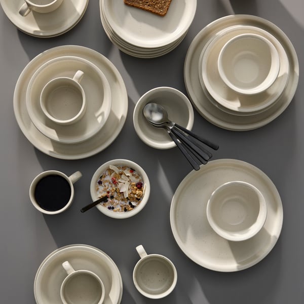 Some DAGGASTER plates, deep plates and bowls are stacked on a grey table together with some cups and spoons.