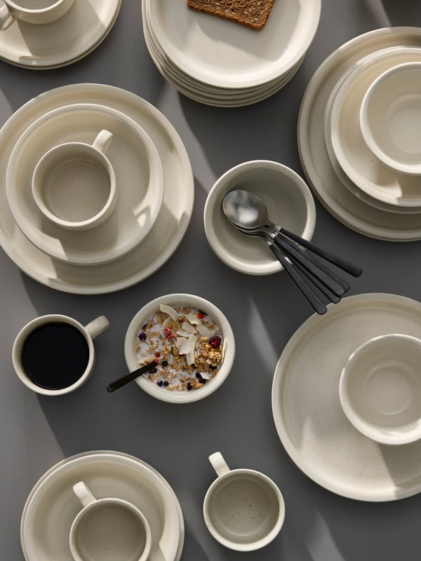 Some DAGGASTER plates, deep plates and bowls are stacked on a grey table together with some cups and spoons.