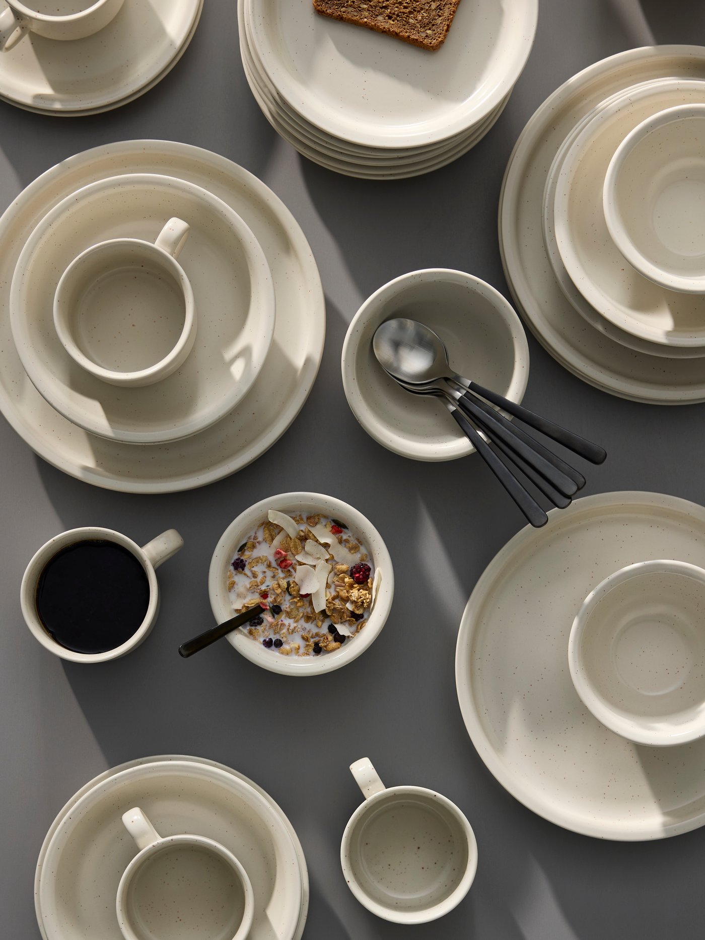 Some DAGGASTER plates, deep plates and bowls are stacked on a grey table together with some cups and spoons.