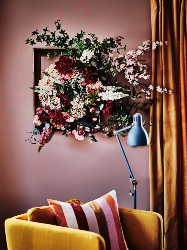 Framed SMYCKA artificial flowers on a wall above a cushion in a pink/striped VATTENVÄN cushion cover on an EKERÖ armchair.