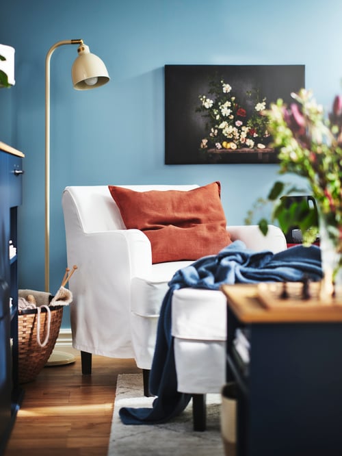 A living room with a RÖDFLIK floor reading lamp, next to an armchair with a red cushion and a picture frame on the wall.