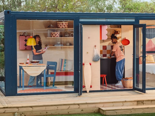 A tiny house on a deck, with glass walls and doors, seen from outside. There are OMMJÄNGE furnishings and decorations inside.