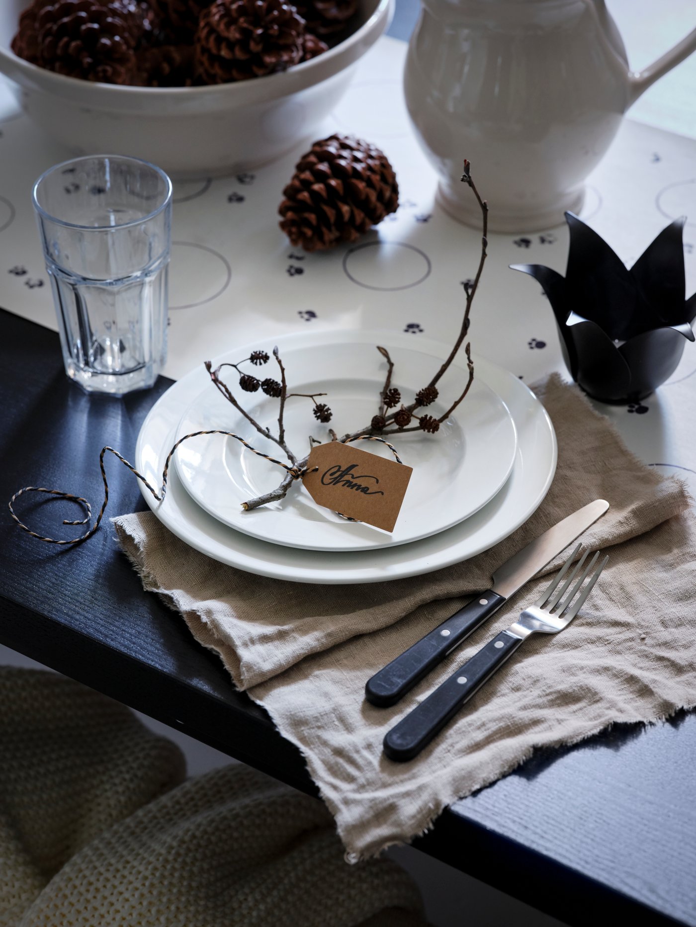 A dinner place setting with two VARDAGEN plates by a POKAL glass and cutlery. Pine cones are in a bowl and on the table.