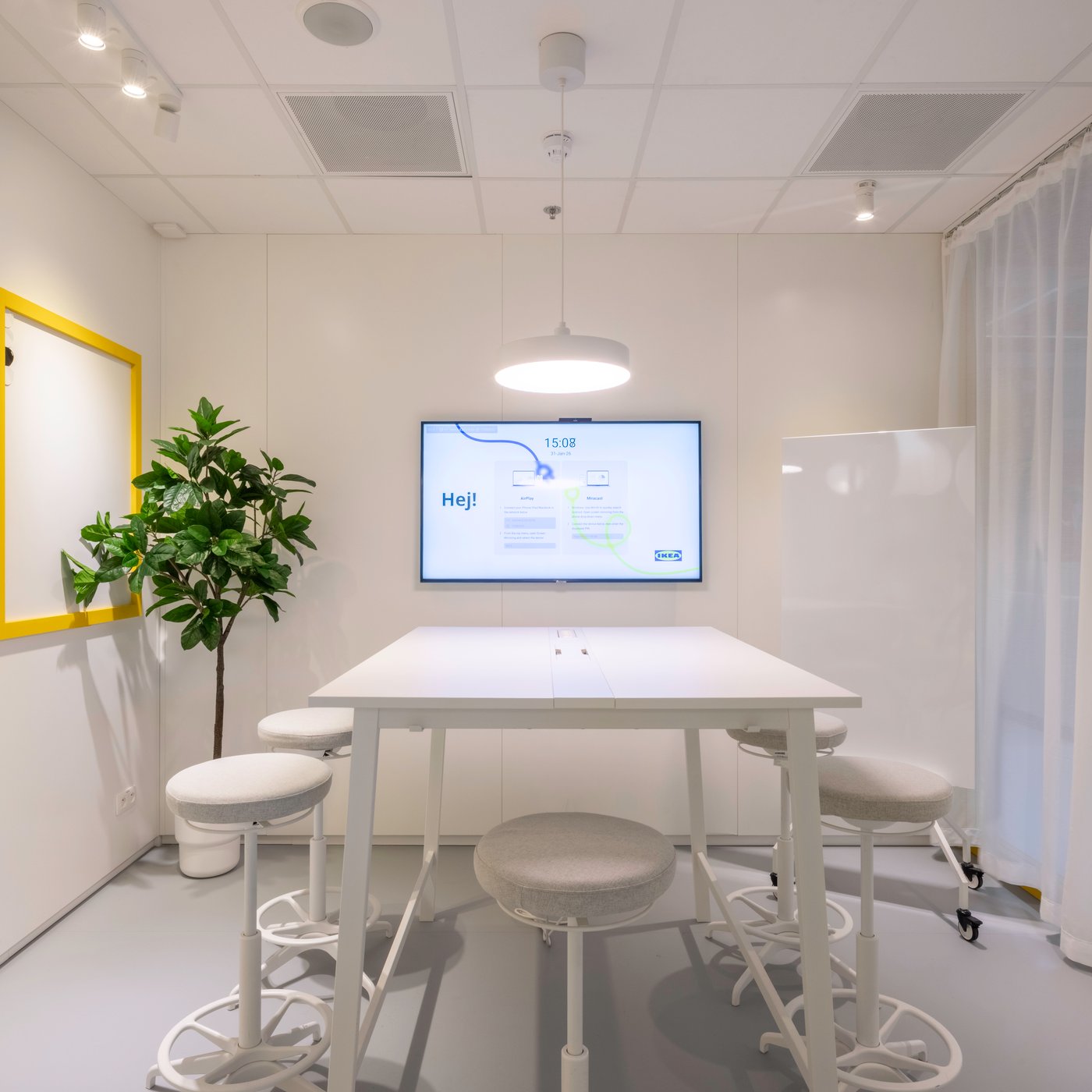 Salle de réunion blanche épurée avec des tabourets ronds, un écran de présentation affichant « Hej ! », un tableau blanc encadré de jaune et une plante verte d’intérieur.