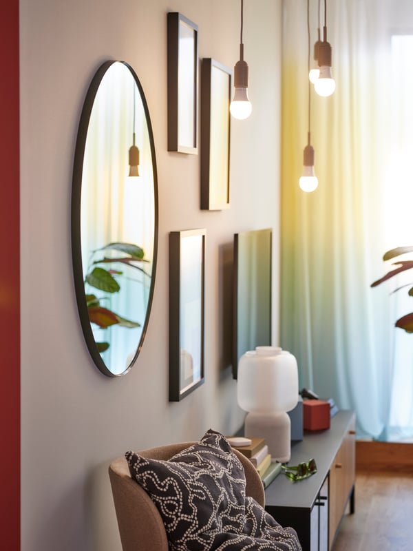 A round LINDBYN mirror, three works of art in RÖDHOLM frames and a TV hang on a wall near some hanging light bulbs.