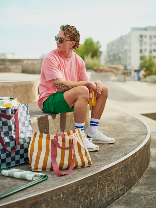 Next to a person holding a carbonated soft drink, there is a SÄCKKÄRRA carrier bag with a cushion inside and a folding stool.