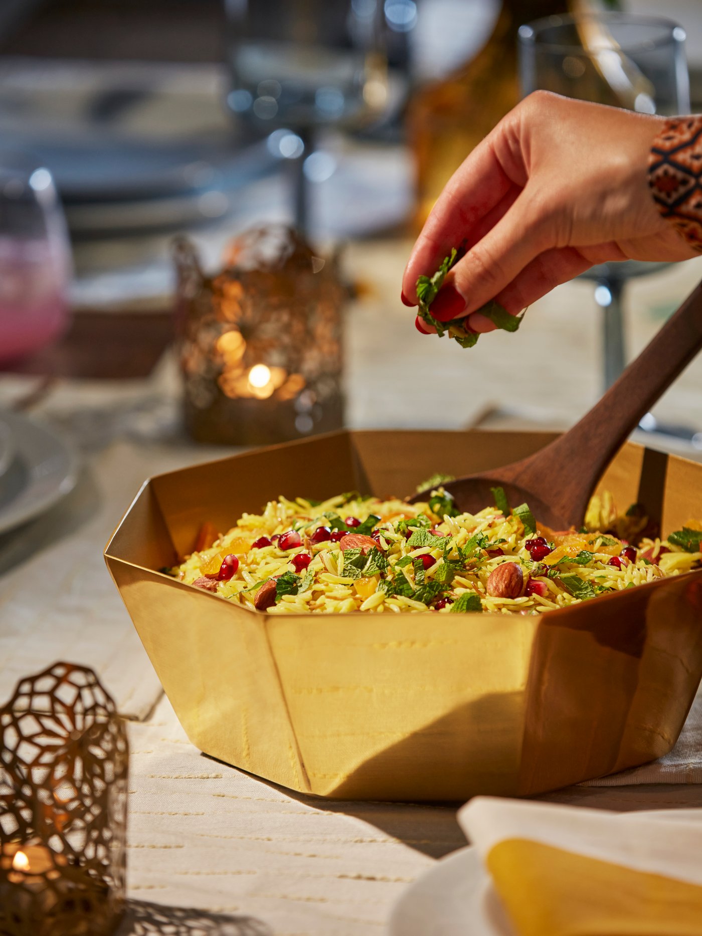 A gold-colour GOKVÄLLÅ serving bowl filled with spiced mediterranean rice. A hand is sprinkling some garnish on the rice.