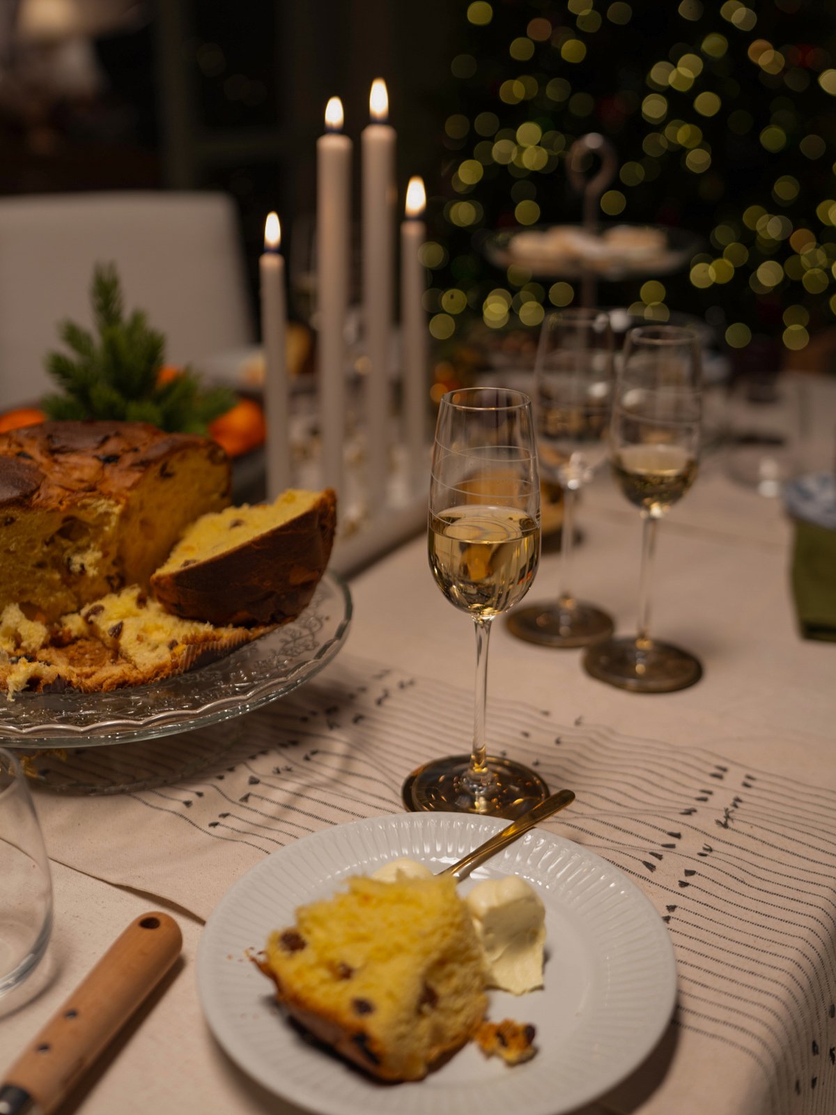 Un dolce panettone a fette su un piatto di vetro, con dietro un candelabro bianco con cinque candele accese e un albero di Natale illuminato.