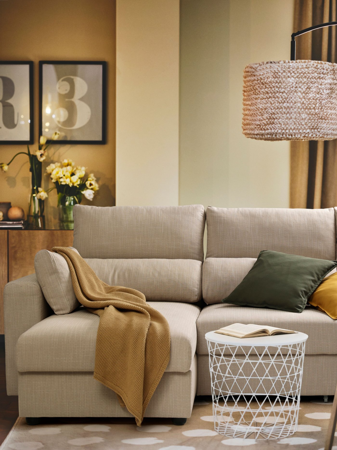 Divano ESKILSTUNA a 3 posti con chaise longue in tessuto beige, in un soggiorno accogliente sui toni caldi. Decorato con plaid senape e cuscini decorativi verdi e senape. 