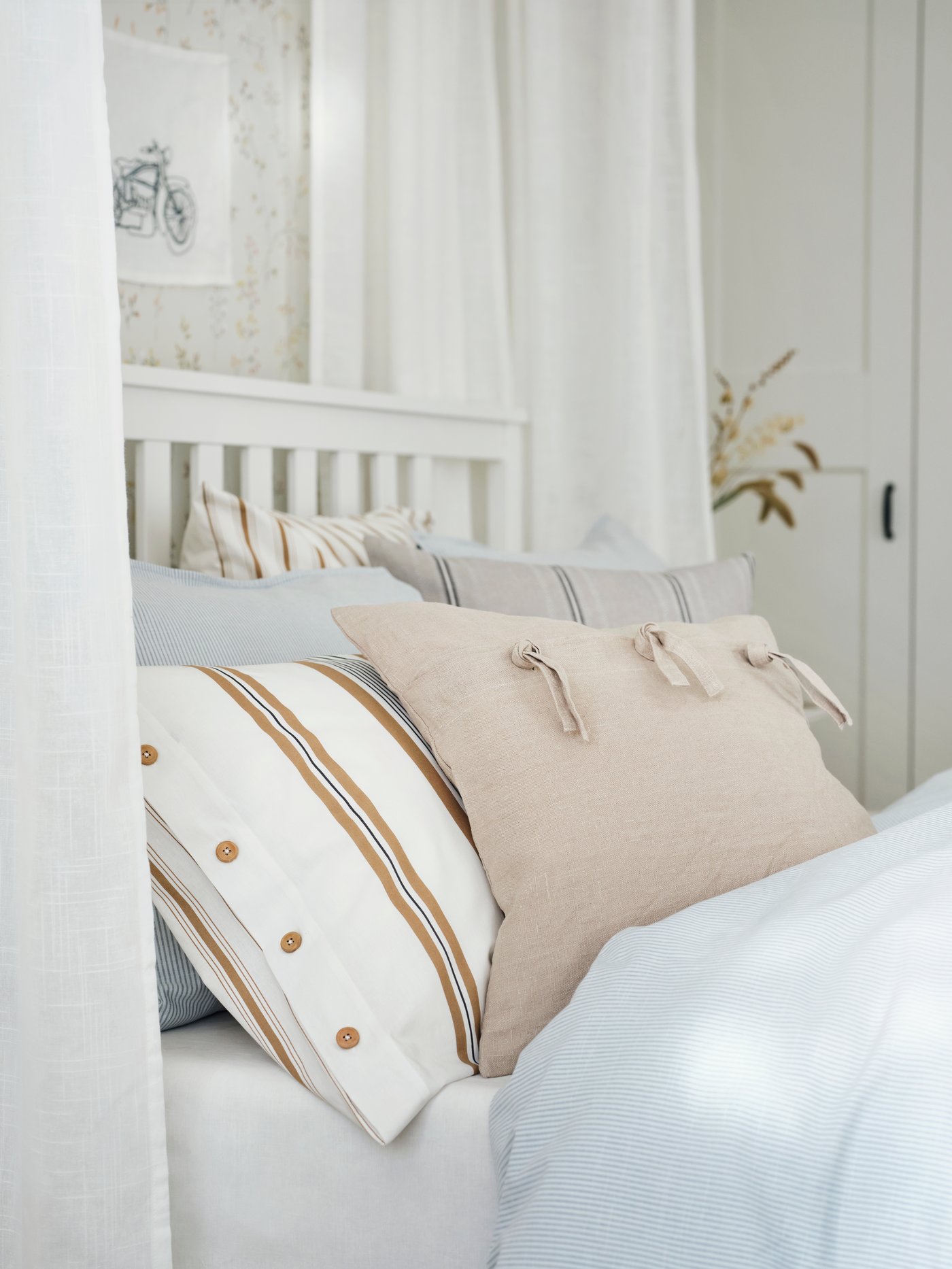 Dormitorio con sábana bajera blanca de algodón con varias fundas de cojín cuadradas y una funda de almohada encima.