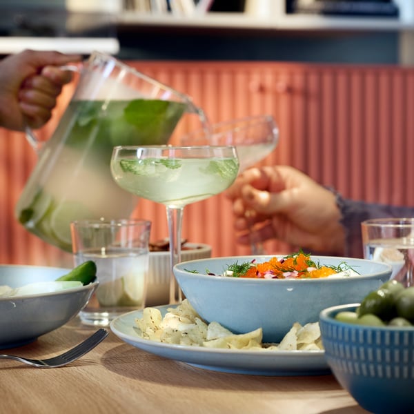 Valaki ünnepi italt tölt egy másik személy poharába, az asztalon pedig világoskék FÄRGKLAR étkészlet látható.