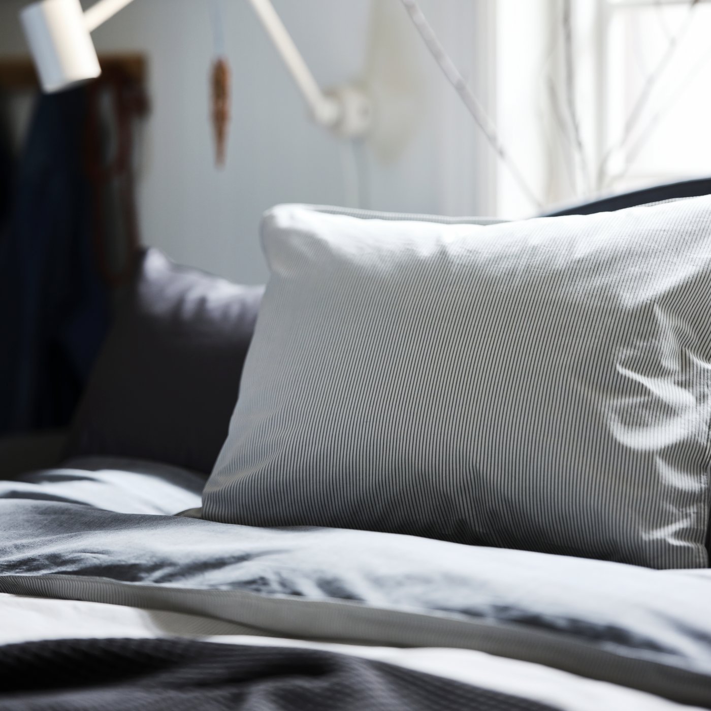 A bedroom with a grey BLÅVINDA quilt and pillowcase on the bed and a white lamp on the wall behind.