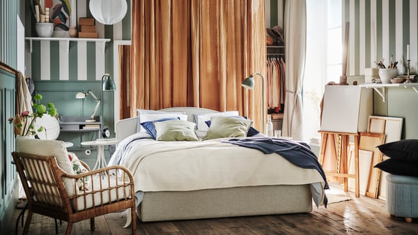 A beige RAMNEFJÄLL upholstered bed in a rustic room with dark beige DYTÅG curtains behind it and a HOLMSTA armchair in front.