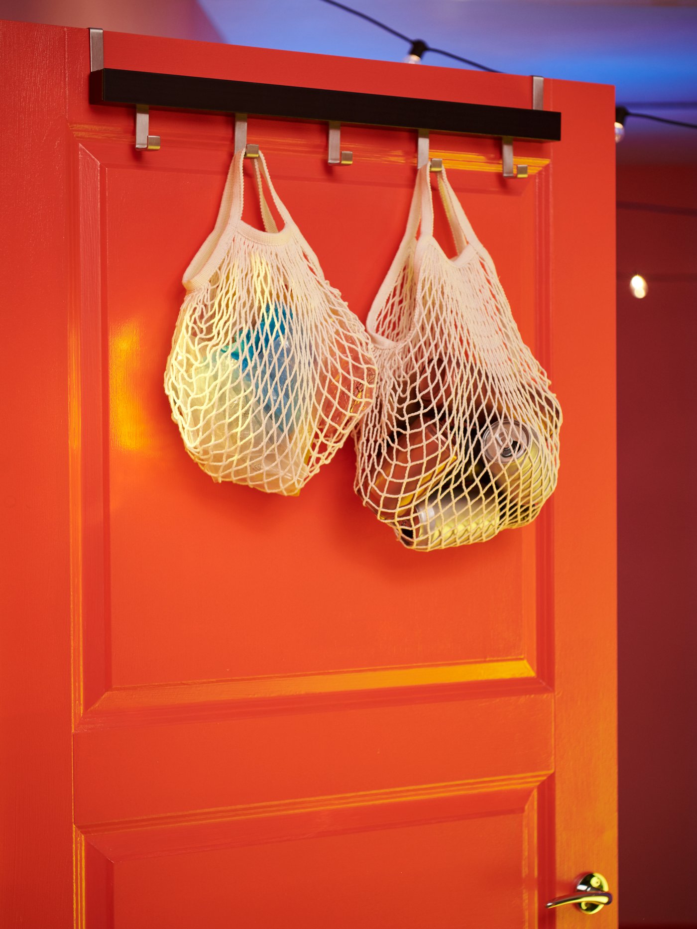 Vestíbulo con un conjunto de dos bolsas de red con objetos, colgadas en una percha negra con cinco pomos, colocadas en una puerta naranja.