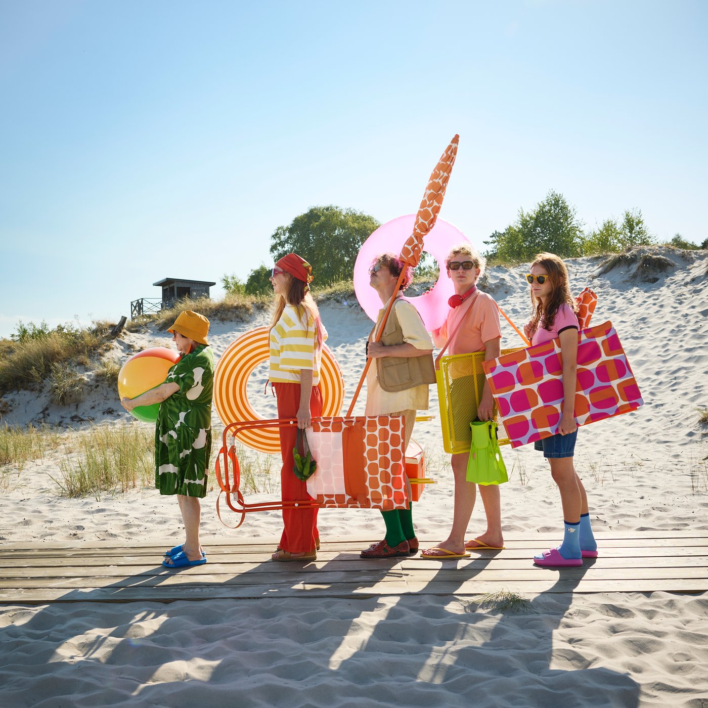 A beach scene with five people holding colourful pool floats and a bright orange STRANDÖN beach chair on the sand.
