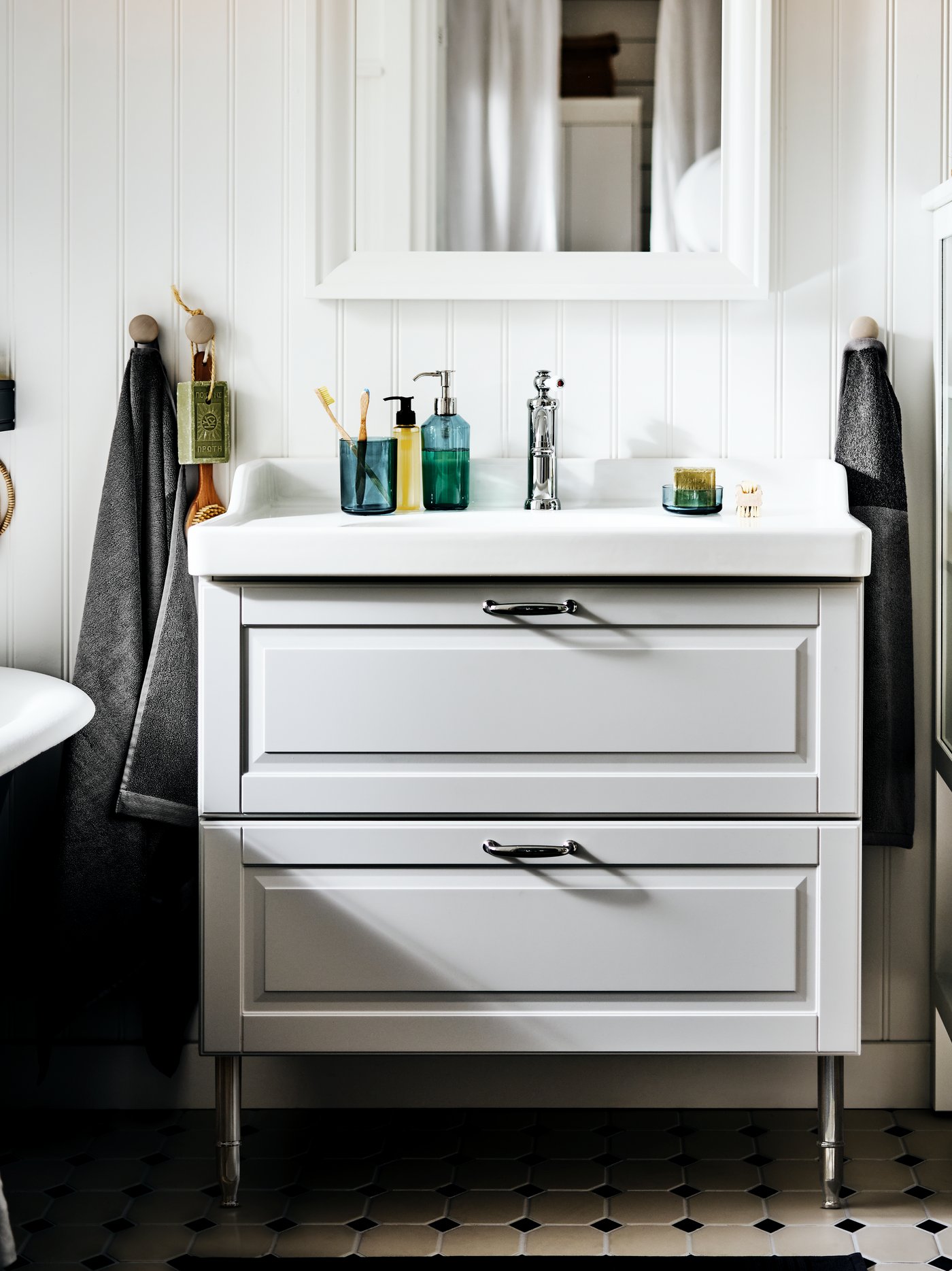 Vista frontal de un vanitorio con lavamanos GODMORGON con dos cajones con canterías y manillas de diseño clásico, de color gris claro, apoyado sobre patas metálicas. Sobre el lavamanos se ve la grifería y accesorios de baño, así como la parte inferior de un espejo TOFTBYN rectangular con marco blanco.