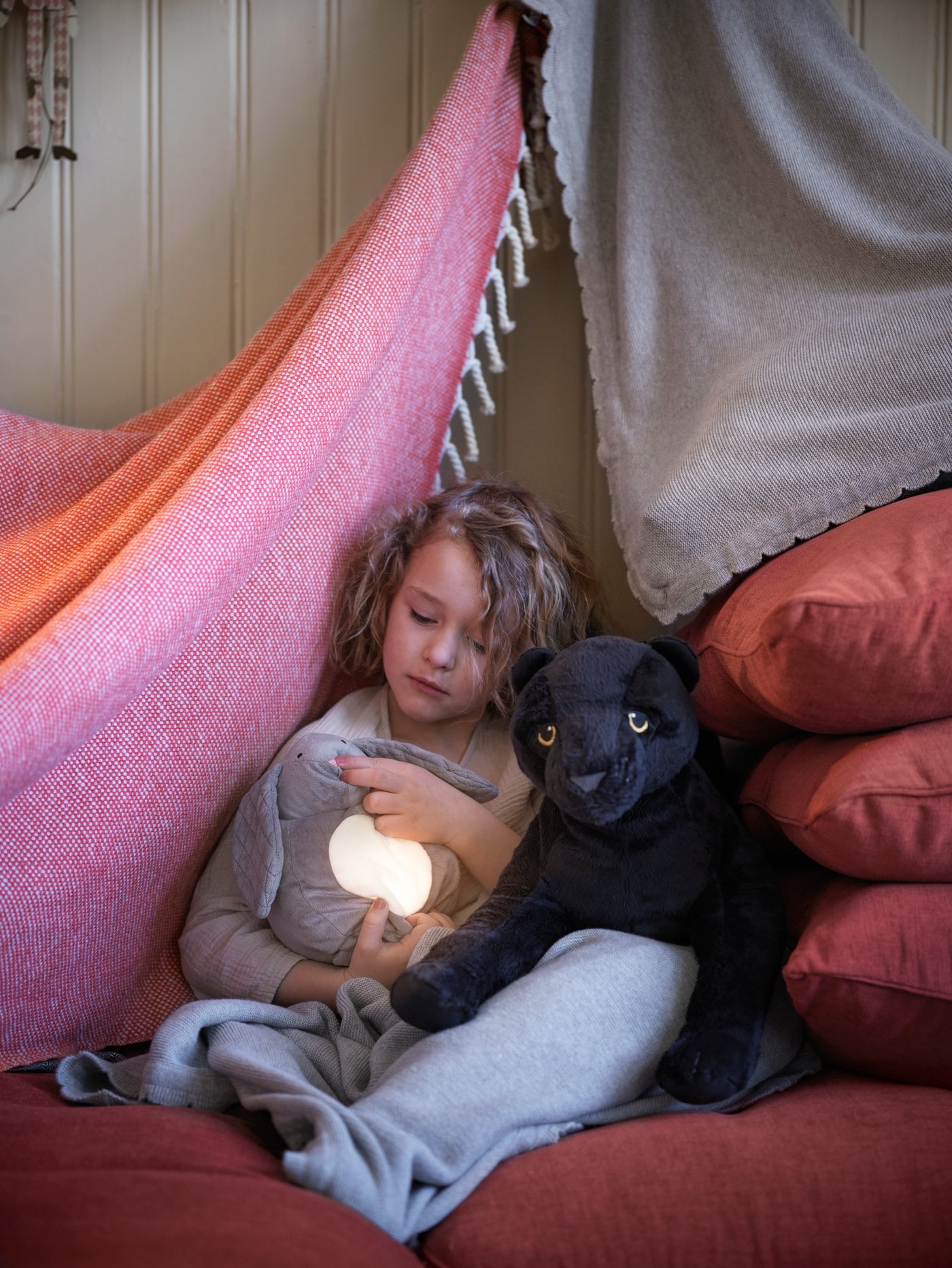 A little girl cradles a grey PEKHULT soft rabbit toy with LED night light in a pink blanket fort, with MJUKHET soft toy.