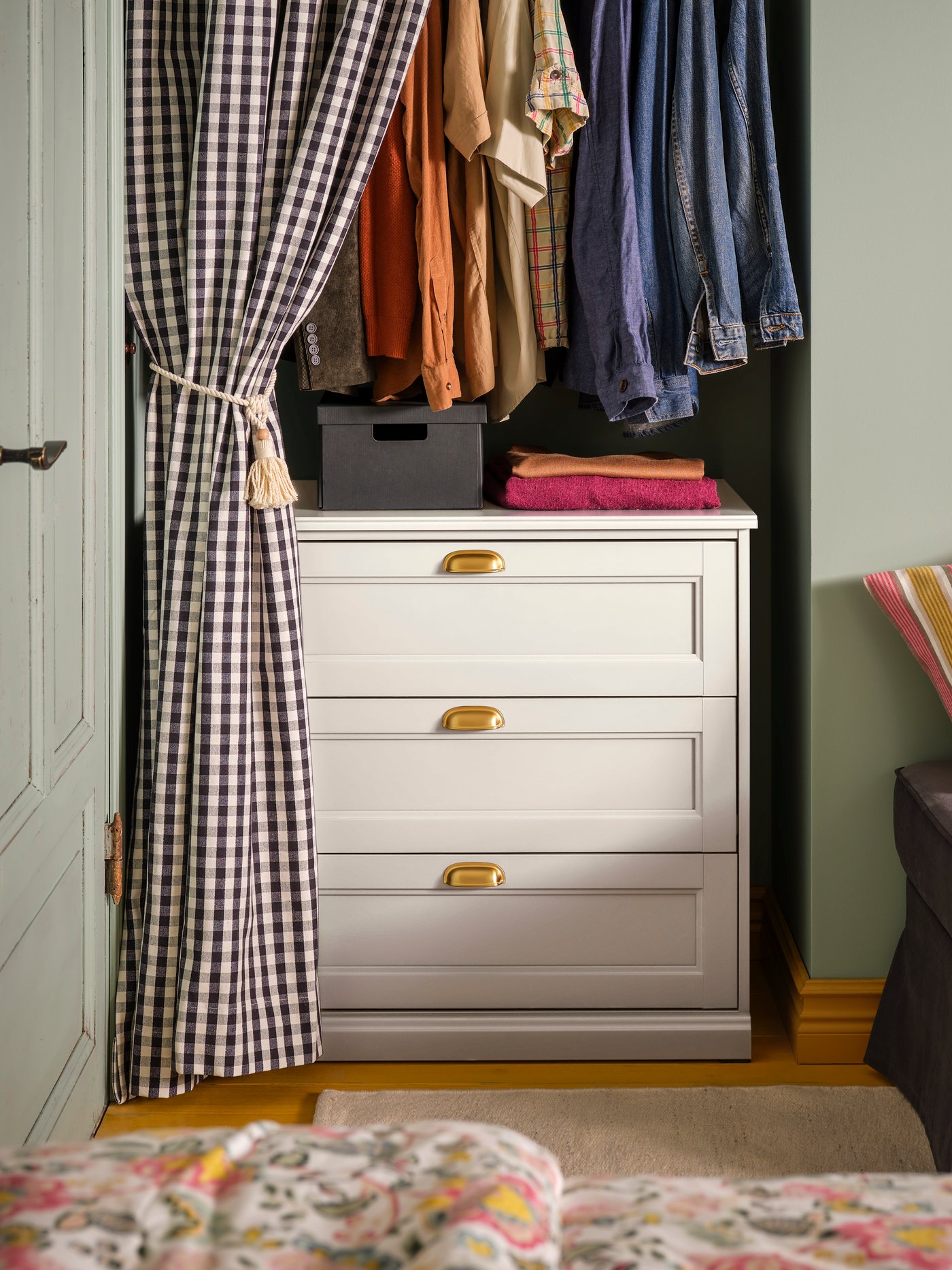 A bedroom with a FÅGELFJÄLLET chest of 3 drawers inside an alcove with clothes hanging above it, behind a checked curtain.