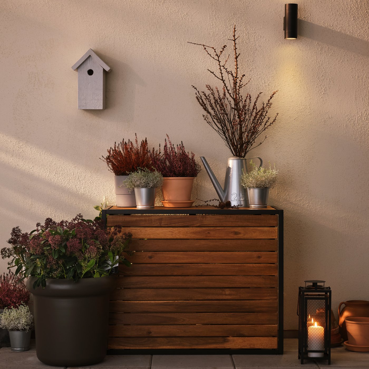 Unha mesa de centro HÅKANSKÄR contra unha parede nunha terraza; sobre a mesa, hai plantas en testos e unha regadeira ÅKERBÄR.