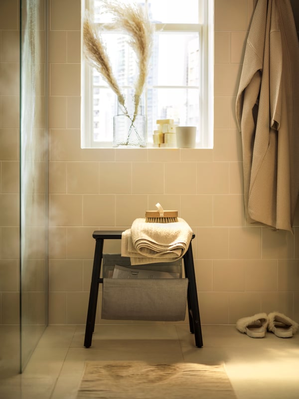Cream tiled bathroom with black storage stool under a window with grasses in rippled glass vase.