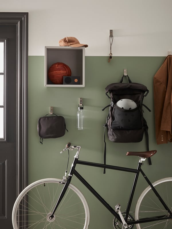 Dark gray VÄRLDENS backpack is hanging on the green wall besides a brown coat and VÄRLDENS toiletry bag. IKEA 365+ water bottle is hanging in the middle of the wall and below EKET wall-mounted shelving unit. A black framed bicycle is parked at the wall near the door.