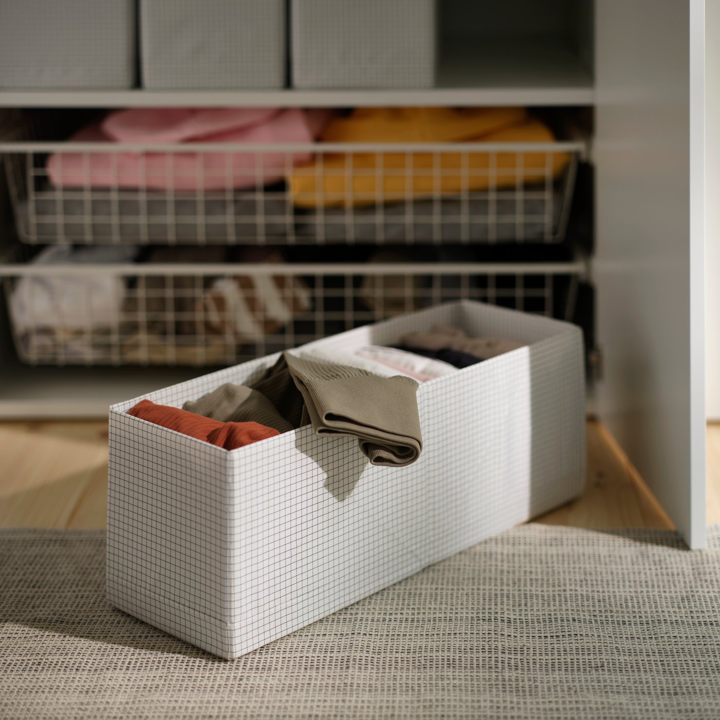 STUK box with compartments in white filled with folded clothes placed on a rug beside wardrobe storage.