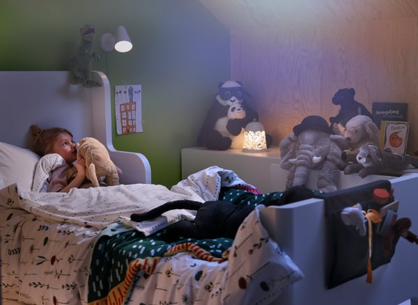 A FUBBLA wall lamp and SOLSKUR table lamp brighten a cozy bedroom as a child in bed whispers to her favorite stuffed toy.