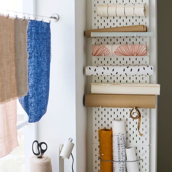Three white SKÅDIS pegboards in a vertical row on a wall with hooks and elastic cord being used to hold rolls of paper.