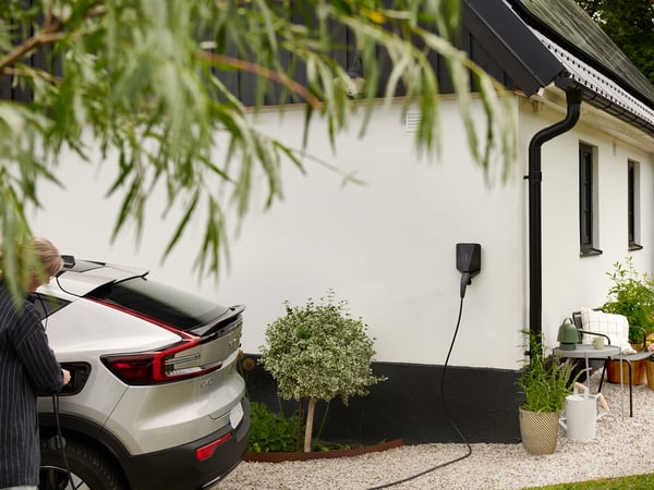 Ein Elektrofahrzeug lädt an einer Wandladestation vor einem modernen Haus, umgeben von Pflanzen und Bäumen.
