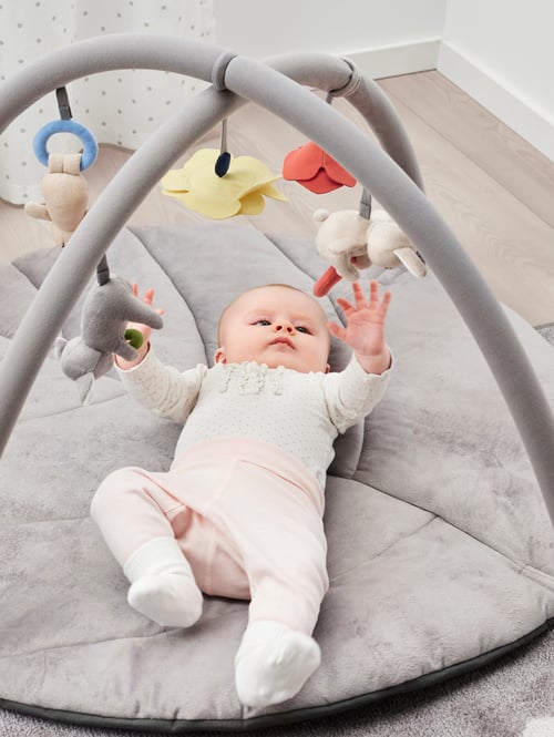 A baby wearing pink trousers and a dotted top lies on its back and plays with the baby toys in a GULLIGAST baby gym.